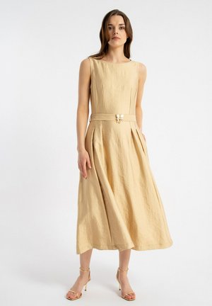 Kobieta w beżowej, bezrękawowej sukience midi z paskiem z motylkowym detalem, w połączeniu z pasującymi sandałami na obcasie, stojąca na białym tle.