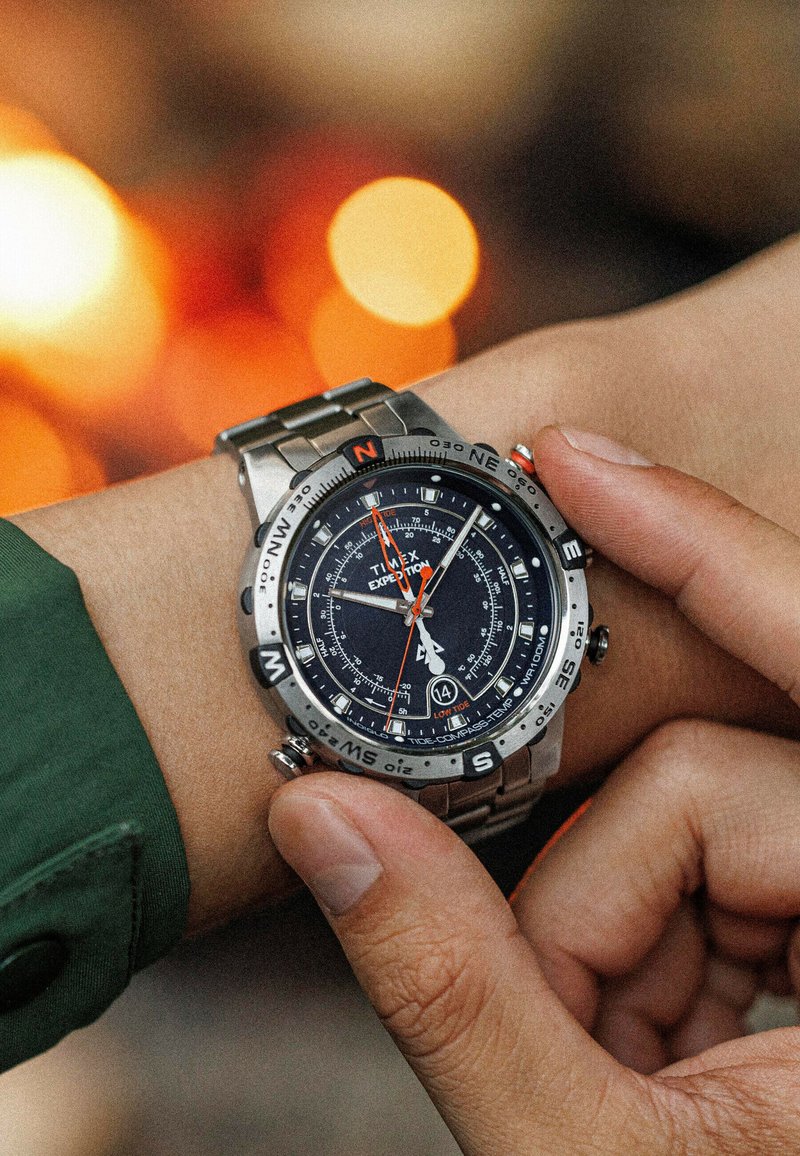 Montre analogique en acier inoxydable avec un cadran noir, des accents orange, des marques de boussole et une lunette texturée. Personne ajustant la montre sur un poignet.