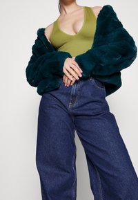 Veste en fausse fourrure verte forêt sur un débardeur côtelé vert olive, assortie à un jean en denim bleu foncé taille haute avec des coutures visibles.