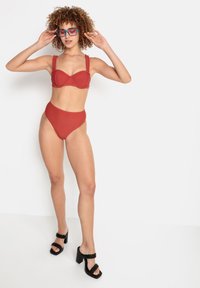 Raudonas dviejų dalių bikinas su struktūruotais puodeliais ir plačiomis petnešėlėmis, derinamas su aukštu liemeniu kelnaitėmis. Juodi platforminiai sandalai su pintomis petnešėlėmis.