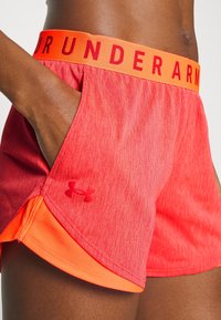Červené sportovní kraťasy s oranžovým pasem, na kterém je nápis "UNDER ARMOUR" tučným písmem. Materiál je hladký s mírným melírovaným vzorem.