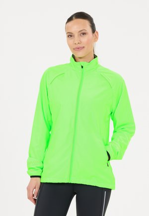 Kvinde iført en lys neongrøn hættejakke med lommer og sorte leggings, poserer mod en ensfarvet hvid baggrund.