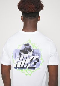 Camiseta de algodón blanca con un gráfico de mancuernas y la palabra "NIKE" en una fuente estilizada, en negrita, negra y gris, con acentos verdes.