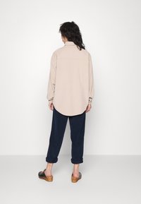 Maglietta oversize beige con bottoni e orlo arrotondato, abbinata a pantaloni blu navy arrotolati e zoccoli con tacco in legno neri. Tessuto texturizzato visibile.