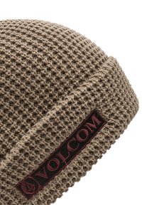 Bonnet en tricot marron en tissu texturé, avec une étiquette noire portant "VOLCOM" en bordeaux. Forme arrondie avec un ajustement snug.
