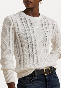 Sweater de cor creme tricotado com padrões em cable, gola redonda, punhos e barra canelados. Usado com calças jeans escuras e um cinto preto com fivela dourada.