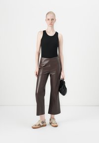 Svart tanktop, brune bukser i faux skinn med bred ben, beige sneakers med mønster, og en liten svart håndveske. Modell står mot en enkel bakgrunn.