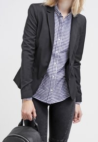 Blazer nero su una camicia a quadri blu e bianca, abbinato a jeans scuri e con un piccolo zaino nero.