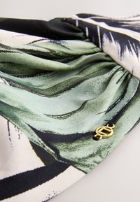 Tissu doux avec des motifs abstraits verts, noirs et blancs, rassemblé avec un petit pendentif ovale doré attaché par un cordon noir.