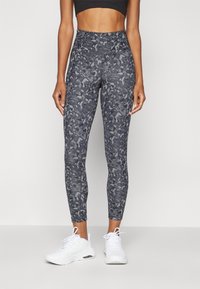 Högmidjade leggings i mörkgrått med camouflagemönster, tillverkade av stretchy material. Bärs med vita träningsskor.