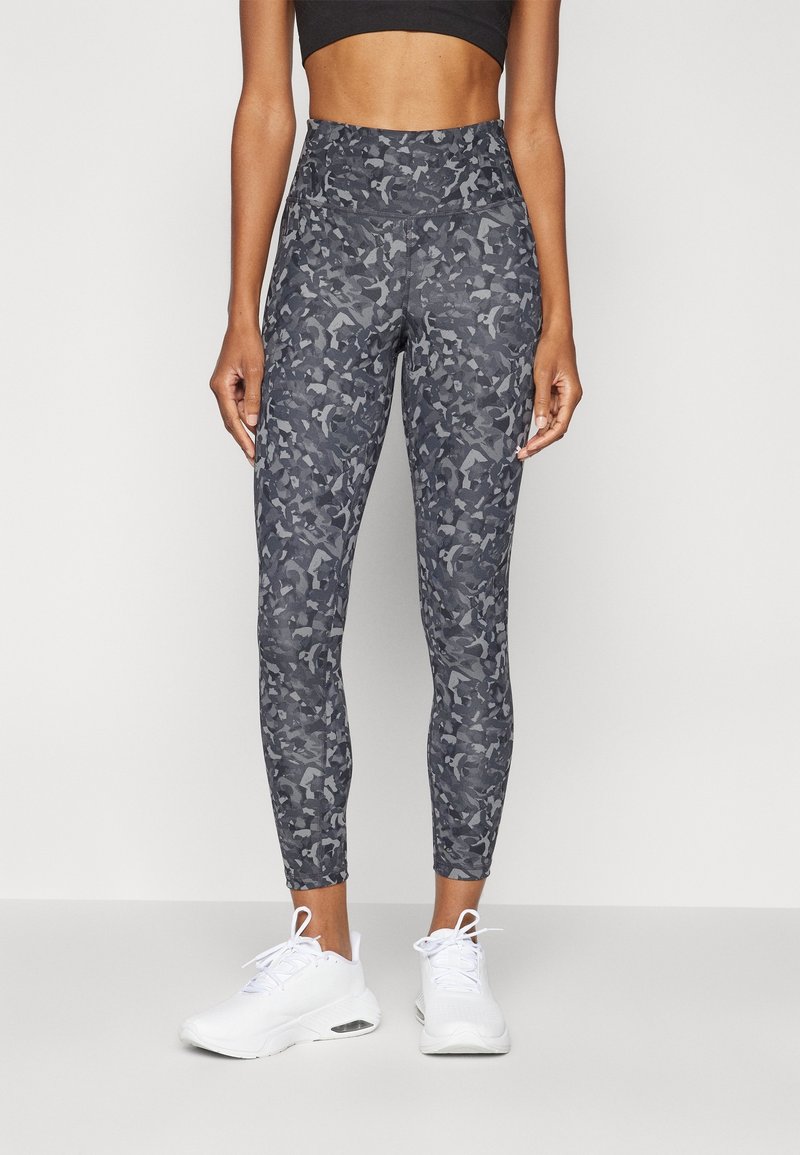 Högmidjade leggings i mörkgrått med camouflagemönster, tillverkade av stretchy material. Bärs med vita träningsskor.