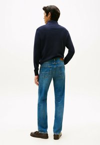 Polo a maniche lunghe blu navy abbinato a jeans dritti azzurri; toppa in pelle sulla cintura; modello di spalle, che indossa mocassini marroni.