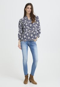 Blouse bleu marine à motif floral blanc, ourlet festonné et manches amples, associée à un jean skinny délavé clair et des bottines marron.