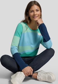 Leichter Pullover in Türkis- und Marineblau-Tönen, mit Wellenmustern. Gerippte Bündchen und eine weiche Textur. Kombiniert mit dunklen Jeans und weißen Sneakers.