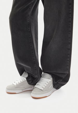 Persona che indossa jeans neri larghi e sneakers Reebok grigio chiaro con lacci bianchi in piedi su una superficie bianca.