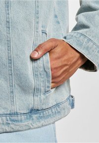 Mano de tono de piel oscura descansando dentro del bolsillo de una chaqueta de denim azul claro, mostrando la textura detallada de la tela y la costura.