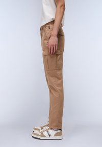 Beige cargo nadrág oldalsó zsebekkel, egyenes szabással és rugalmas derékkal; bézs és fehér sneakers fekete részletekkel párosítva.