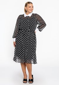 Schwarzes Kleid mit Punkten aus transparentem Stoff, mit weißem Kragen, langen Ärmeln, tailliertem Gürtel und knielangem Saum. Sichtbare schwarze Sandalen mit offenem Zehenbereich.