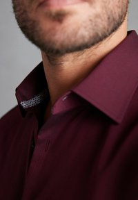 Bordeauxfarbene Hemd aus strukturiertem Stoff mit einem kontrastierenden gemusterten Kragen. Sichtbare Knöpfe am Ausschnitt, die ein modernes Design zeigen.
