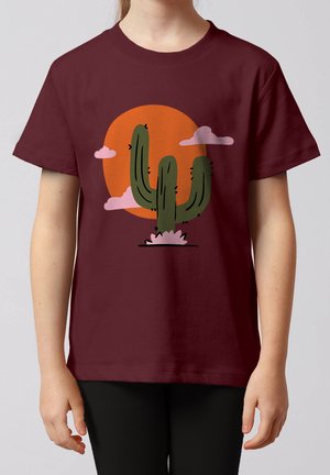 Kind draagt een kastanjebruin t-shirt met een afbeelding van een groene cactus, roze wolken en een oranje zon op de achtergrond.