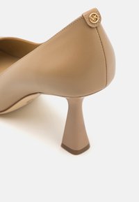 Chaussure à talon haut en cuir beige avec un talon triangulaire unique, texture lisse et accent logo doré à l'arrière du talon.