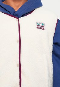Fleece vest, cream color with maroon edging, featuring a blue hooded sweatshirt underneath. Notable tag reads "OVC On Vacation Club."