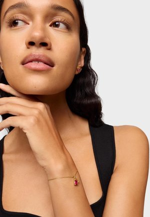 Pulsera de oro con un encantador de gema rosa, llevada en una muñeca. El modelo tiene el cabello oscuro y viste una camiseta sin mangas negra, con la mano descansando cerca de la barbilla.