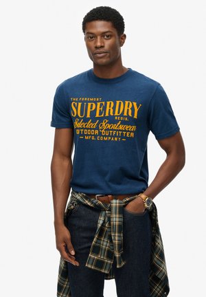Navy blue cotton t-shirt with yellow graphic text. Worn with dark jeans and a green plaid shirt tied at the waist. Watch on wrist.