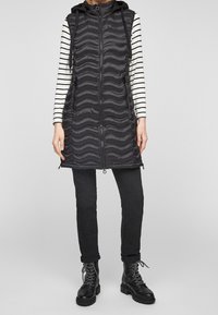 Schwarze Puffer-Weste mit wellenförmiger Steppstruktur, Reißverschluss und Kapuze. Kombiniert mit einem gestreiften Langarmshirt und schwarzen Hosen, dazu robuste Stiefel.