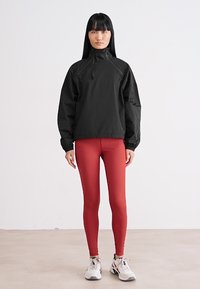 Schwarze, leichte Pulloverjacke mit hohem Kragen und elastischen Bündchen; kombiniert mit figurbetonten roten Leggings und weißen Sportschuhen.