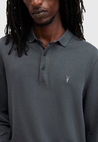 Man met een donkere huid en dreadlocks, gekleed in een donkergrey poloshirt met lange mouwen en een klein geborduurd dierenkoplogo op de borst.
