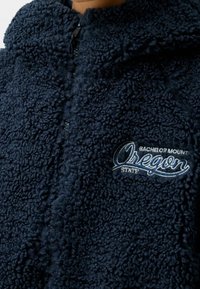 Marintröja i fleece med dragkedja, texturerad tyg, hög krage och broderad "Oregon State Bachelor Mount"-logotyp i ljusblått.
