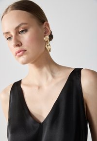 Orecchini astratti dorati con finitura lucida, indossati con un top senza maniche nero caratterizzato da una profonda scollatura a V, texture liscia e design minimal.