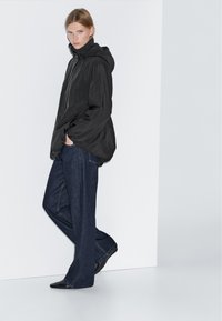 Donna con giacca a cappuccio nera, jeans larghi di un blu scuro e scarpe nere a punta, in piedi contro un muro bianco con le mani in tasca.