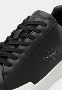 Μαύρο δερμάτινο αθλητικό παπούτσι με υφή, που διαθέτει μαύρα κορδόνια, ανάγλυφο λογότυπο και αντίθετη λευκή ελαστική σόλα.