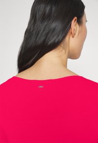 Maglione aderente a coste di un vivace colore rosa. Presenta un ampio scollo e una piccola etichetta del marchio sul retro. Texture liscia e maniche lunghe.