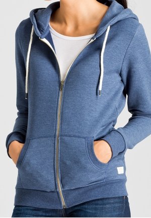 Osoba ubrana w niebieską bluzę z zamkiem na suwak i białymi sznurkami, na białej koszulce, ręce w kieszeniach i ciemnoniebieskie dżinsy.