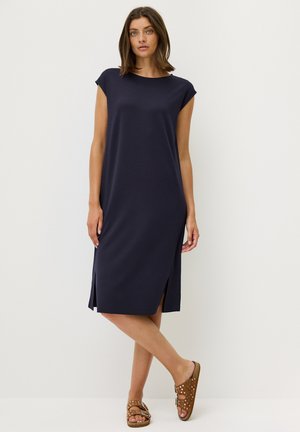Robe bleu marine, longueur genoux, avec de courtes manches à capuchon, fentes latérales et une coupe décontractée, fabriquée en tissu doux. Portée avec des sandales marron.