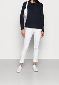 Vrouw in een marineblauwe lange mouwen top, witte opgerolde jeans, witte sneakers en met een witte schoudertas op een lichte achtergrond.