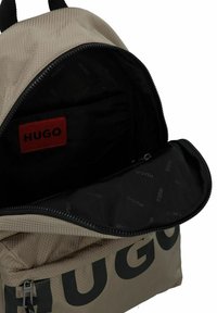Mochila beis con un exterior texturizado, un gran logo negro y una apertura con cremallera que revela un interior negro con una etiqueta de logo roja.