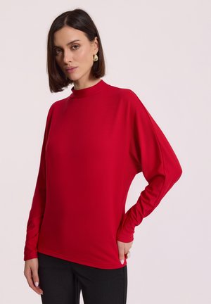 Kobieta o średniej długości brązowych włosach, ubrana w czerwoną bluzkę z długimi rękawami i czarne spodnie, stojąca na tle jasnym jednolitym.