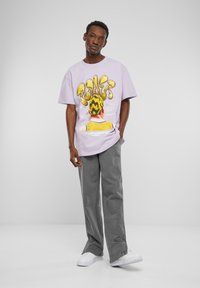 Lilla oversized t-shirt med grafisk tekst og en illustration på ryggen, parret med grå brede bukser og hvide sneakers.