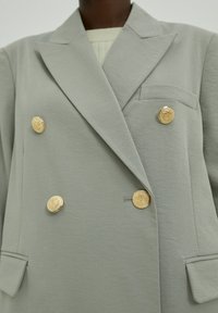 Blazer gris croisé en tissu doux, avec des boutons décoratifs dorés et une poche poitrine. Lignes épurées et design structuré.
