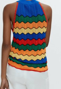 Vrouw in een mouwloze gebreide top met meerkleurige golvende strepen in blauw, groen, oranje, geel en rood, gecombineerd met witte broek.