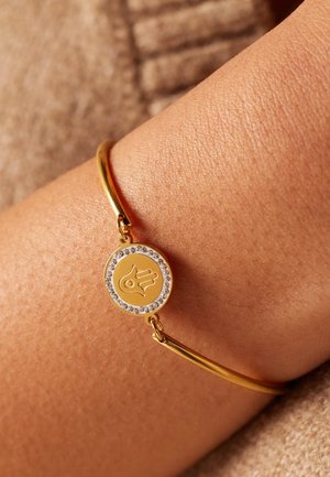 Goldarmband mit einem runden Anhänger, der ein Handdesign zeigt, umgeben von klaren Strasssteinen, auf einem glatten, glänzenden Band.