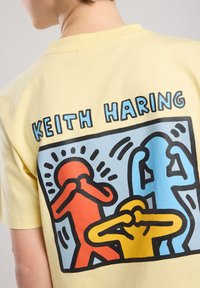 Keltteä puuvillapaita, jossa on Keith Haringin graafinen suunnittelu, esittää kolmea abstraktia hahmoa punaisissa, keltaisissa ja sinisissä ääriviivoissa.