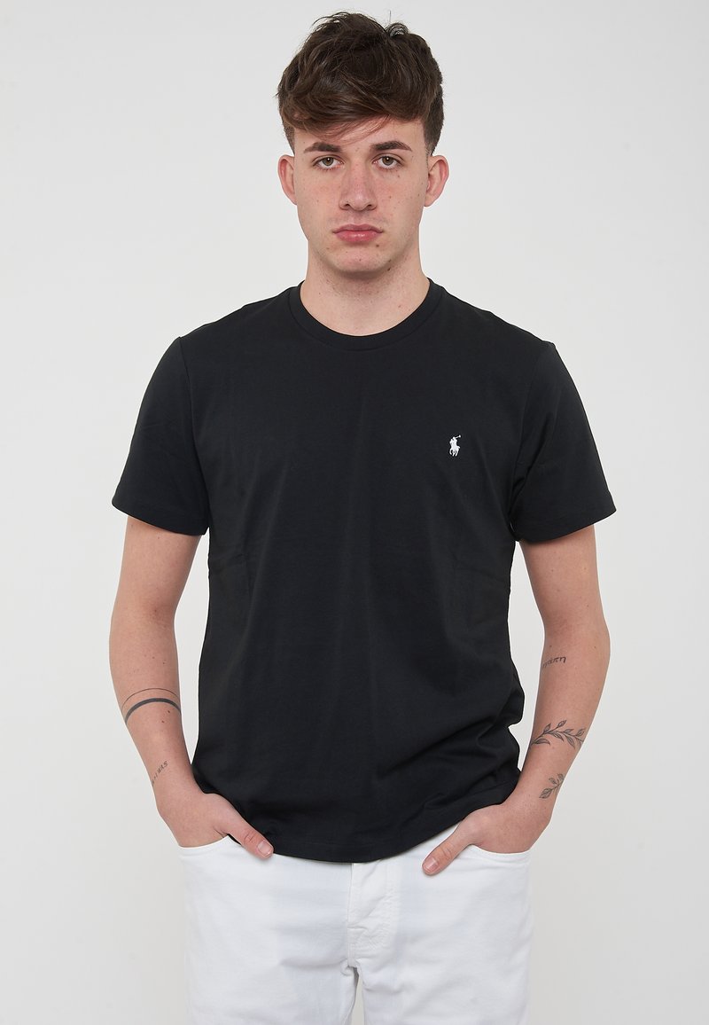 T-shirt in cotone nero con scollatura rotonda, maniche corte e un piccolo logo bianco sul lato sinistro del petto. Indossato con pantaloni bianchi.