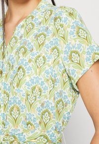 Camicia floreale a maniche corte di colore verde chiaro con dettagli blu e oro. Presenta un motivo a ripetizione di fiori e foglie, con una tasca decorativa.