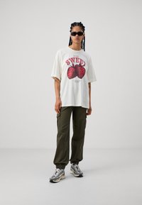 Cremefarvet t-shirt med et rødt jordbærgrafik, parret med olivengrønne cargo-bukser. Modellen har sorte solbriller og mønstrede sneakers på.
