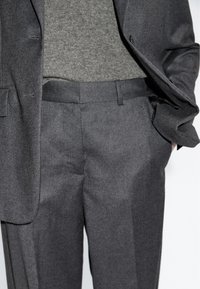 Grau maßgeschneiderter Anzug mit passenden grauen Hosen, der eine glatte Textur und klare Linien aufweist. Ein grauer Strickpullover wird darunter getragen.